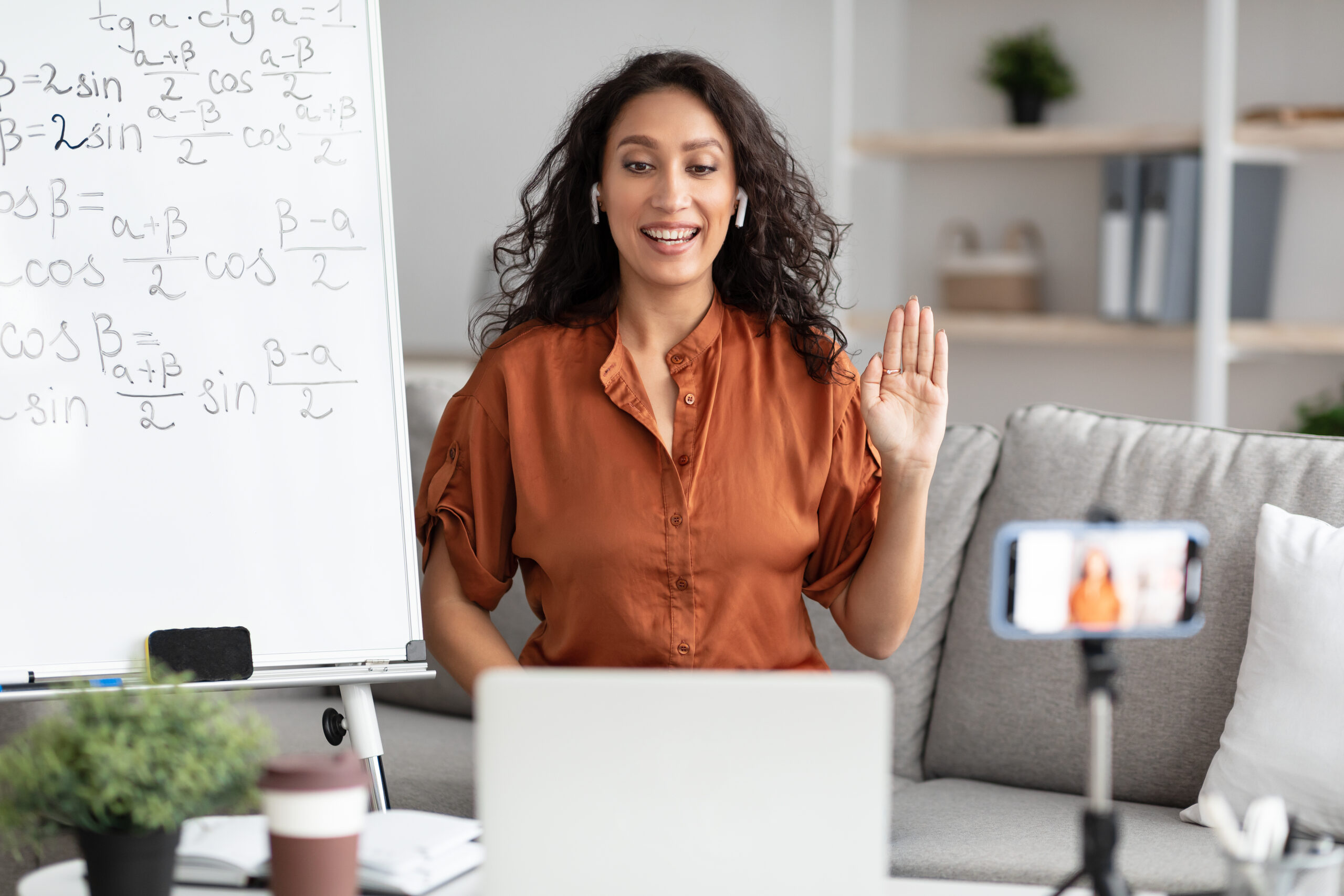 Online Education. Happy female teacher in wireless earbuds having virtual meeting with students on laptop, sitting at desk, waving hand to webcam shooting video training on cell phone using tripod